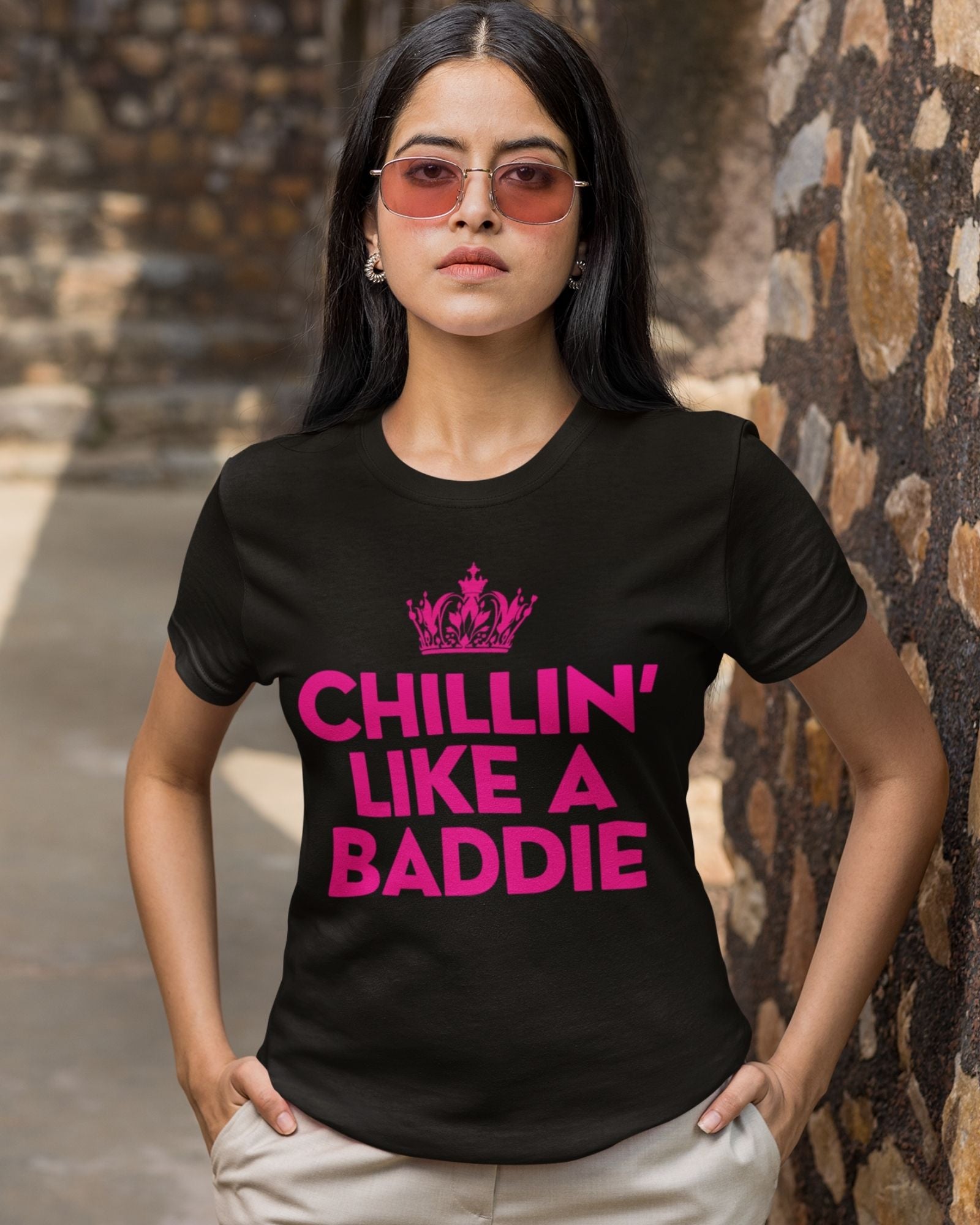 Woman wearing a black t-shirt with pink text and sunglasses, standing against a stone wall.