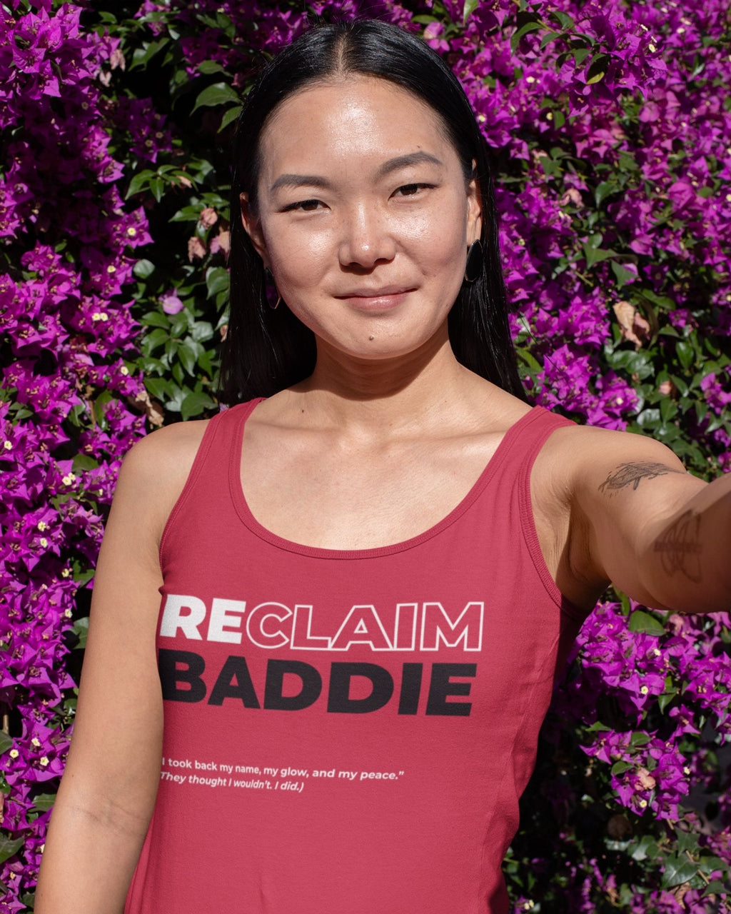 Woman wearing a red tank top with 'RECLAIM BADDIE' text, standing in front of purple flowers.
