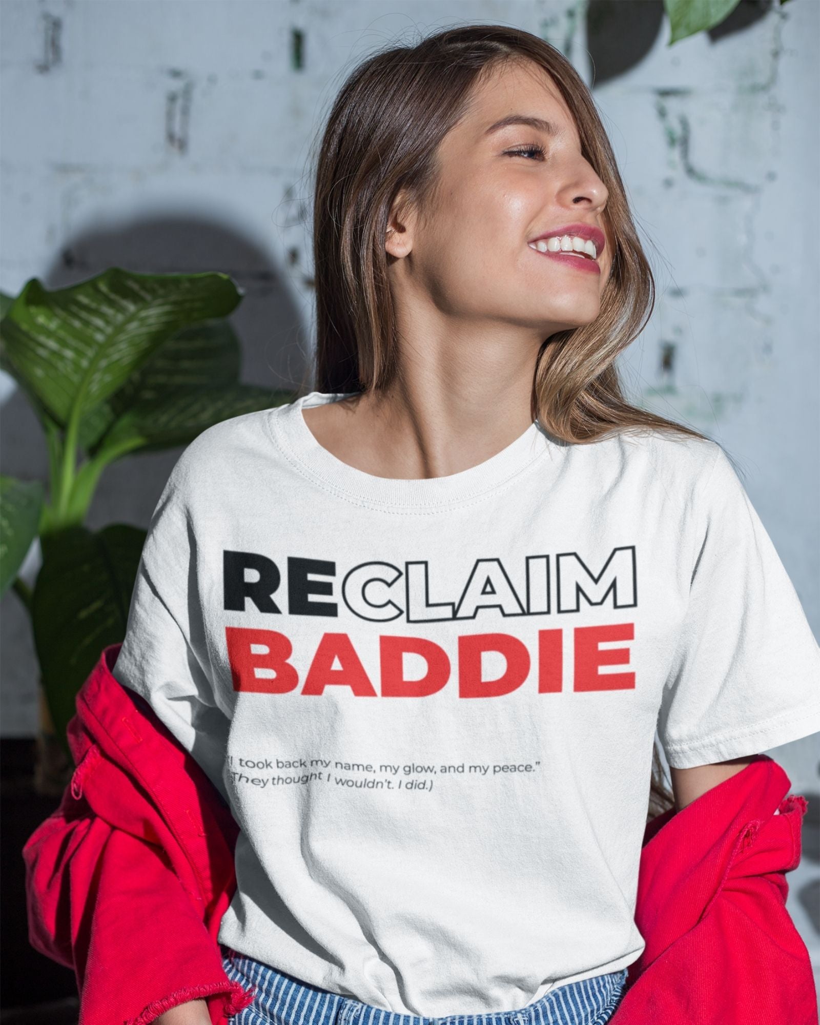Woman wearing a white t-shirt with 'RECLAIM BADDIE' text, standing indoors with plants in the background.